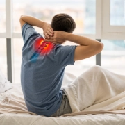 hombre levantándose de la cama con dolor cervical, y esa zona de la espalda marcada en rojo como símbolo de dolor.